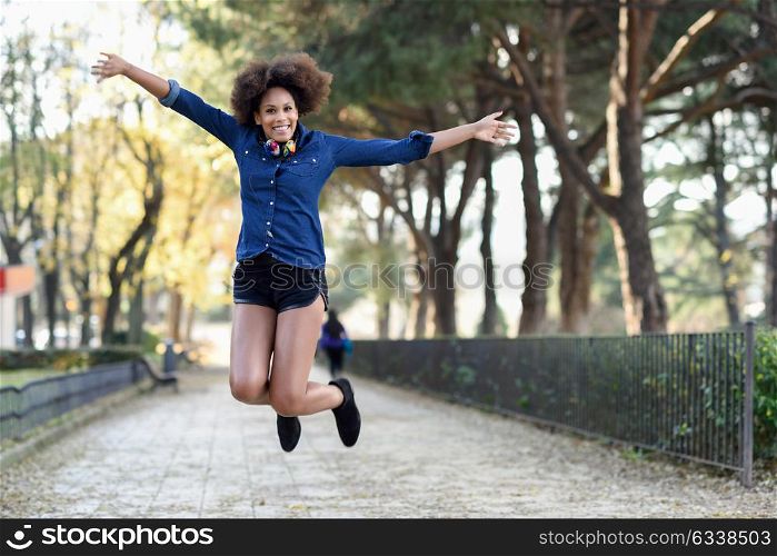 Young black woman with afro hairstyle jumping in urban background with open arms. Mixed woman wearing blue shirt and shorts. Female carrying funny headphones.