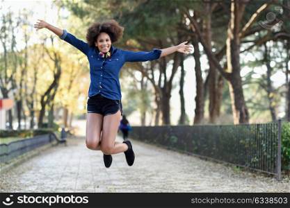 Young black woman with afro hairstyle jumping in urban background with open arms. Mixed woman wearing blue shirt and shorts. Female carrying funny headphones.