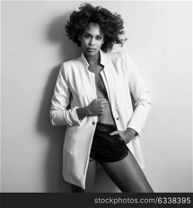 Young black woman with afro hairstyle possing near a white wall in her room. Mixed girl wearing blazer jacket and shorts.