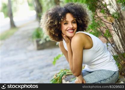 Young black woman with afro hairstyle sitting and smiling in urban park. Mixed girl wearing white t-shirt and blue jeans.