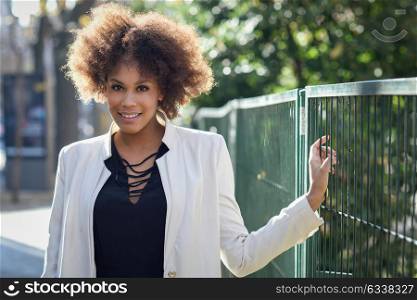 Young black woman with afro hairstyle smiling in an urban street. Mixed girl wearing white jacket and black dress with city background.