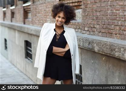 Young black woman with afro hairstyle smiling in urban background. Mixed girl wearing white jacket and black dress posing near a brick wall