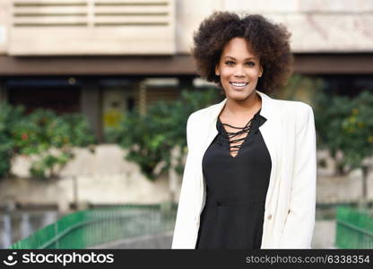 Young black woman with afro hairstyle smiling in urban background. Mixed girl wearing white jacket and black dress.