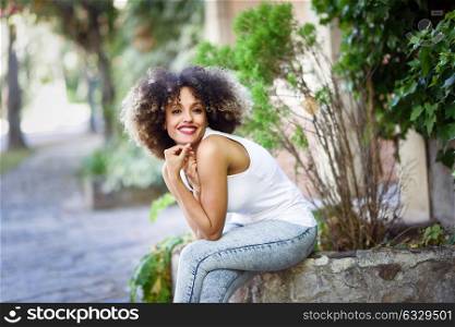 Young black woman with afro hairstyle smiling in urban park. Mixed girl wearing casual clothes sitting in the street.