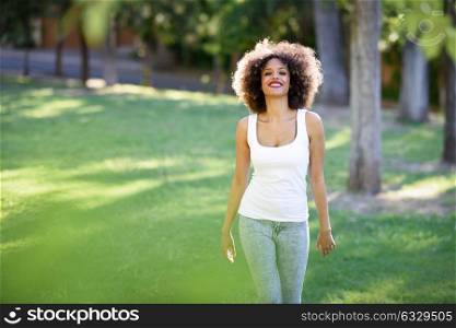 Young black woman with afro hairstyle smiling in urban park. Mixed girl wearing white t-shirt and blue jeans walking on the grass.