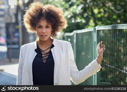 Young black woman with afro hairstyle standing in an urban street. Mixed girl wearing white jacket and black dress with city background.