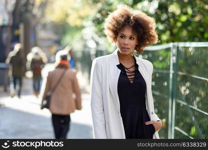 Young black woman with afro hairstyle standing in an urban street. Mixed girl wearing white jacket and black dress with city background.