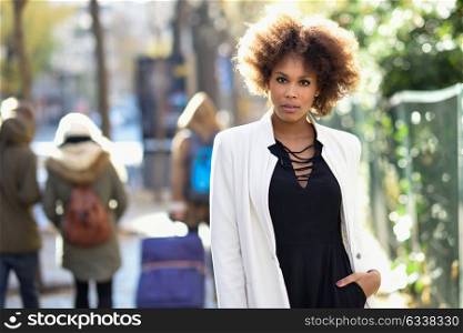 Young black woman with afro hairstyle standing in an urban street. Mixed girl wearing white jacket and black dress with city background.