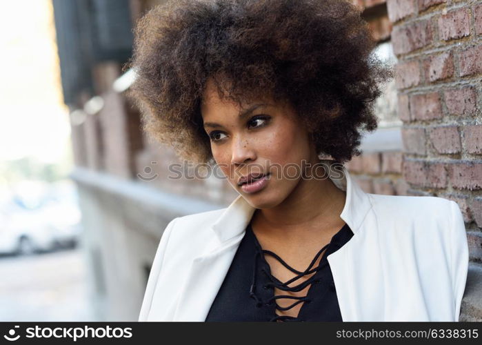 Young black woman with afro hairstyle standing in urban background. Mixed girl wearing white jacket and black dress posing near a brick wall