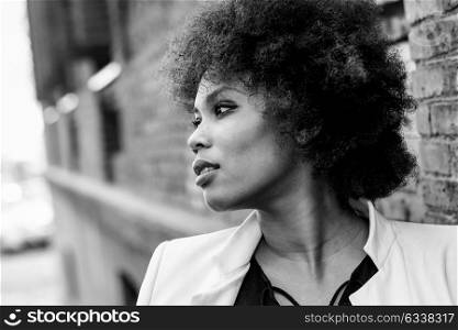 Young black woman with afro hairstyle standing in urban background. Mixed girl wearing white jacket and black dress posing near a brick wall