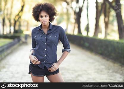 Young black woman with afro hairstyle standing in urban background. Mixed woman wearing blue shirt and shorts.