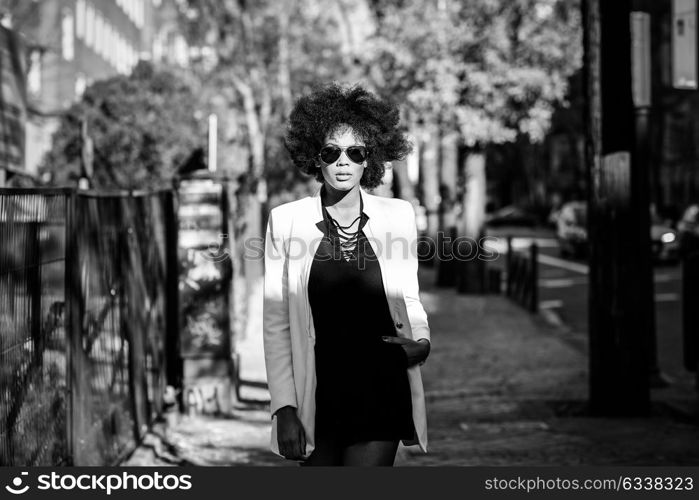 Young black woman with afro hairstyle standing in urban background with aviator sunglasses. Mixed girl wearing white jacket and black dress posing near a brick wall