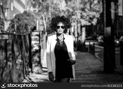 Young black woman with afro hairstyle standing in urban background with aviator sunglasses. Mixed girl wearing white jacket and black dress posing near a brick wall