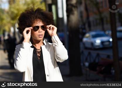 Young black woman with afro hairstyle standing in urban background with aviator sunglasses. Mixed girl wearing white jacket and black dress posing near a brick wall