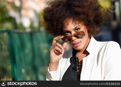 Young black woman with afro hairstyle standing in urban background with aviator sunglasses. Mixed girl wearing white jacket and black dress posing near a brick wall