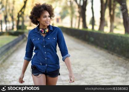 Young black woman with afro hairstyle walking in urban background. Mixed woman wearing blue shirt and shorts. Female carrying funny headphones.