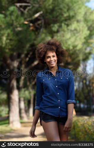Young black woman with afro hairstyle walking in urban park. Mixed woman wearing blue shirt and shorts. Female smiling.