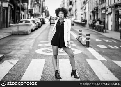 Young black woman with afro hairstyle walking on a crosswalk in an urban street. Mixed girl wearing white jacket and black dress with city background.