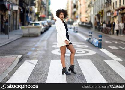 Young black woman with afro hairstyle walking on a crosswalk in an urban street. Mixed girl wearing white jacket and black dress with city background.