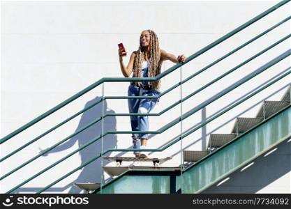Young black woman with coloured braids, looking at her smartphone with her feet resting on a skateboard.. Black woman with coloured braids, looking at her smartphone with her feet resting on a skateboard.