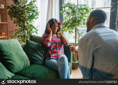 Young black woman with red rose sitting on sofa, romantic meeting at home. Happy african american female person on the couch in living room. Young black woman with red rose sitting on sofa