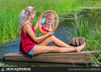 Young blond caucasian woman sitting with mirror at water in free nature