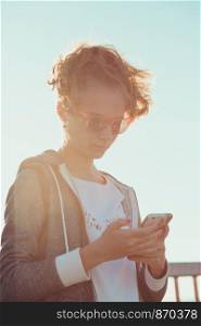 Young blond hair girl using phone, looking at screen, standing outdoors, she is backlighted by sunlight with plain sky in the background