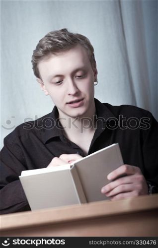 Young blond man looking into open book sitting at table.. Young man reading a book