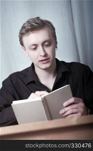 Young blond man looking into open book sitting at table.. Young man reading a book