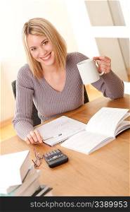 Young blond student writing homework and holding cup of coffee