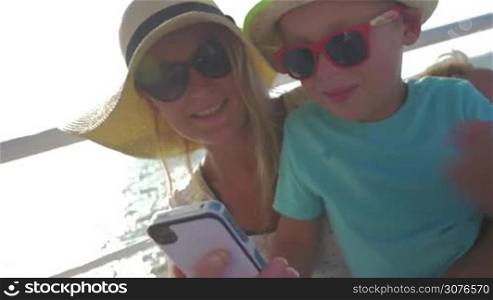 young blond woman and her small son in straw hat and sun glasses are watching something in smartphone against riding on water in boat background summer time.