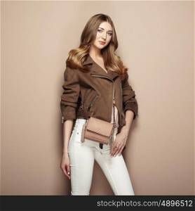 Young blond woman in brown jacket and white jeans. Girl posing on a beige background. Jewelry and hairstyle. Girl with brown handbag. Fashion photo