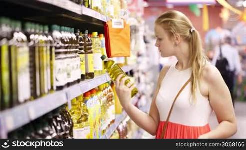 Young blond woman picking an olive oil bottle from the shelves of a supermarket and reading the label