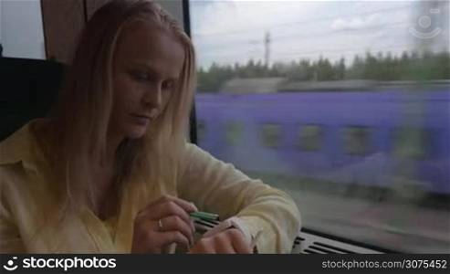 Young blond woman sitting by the window in train and using smart watch with pen