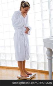 young blond woman wearing a white bathrobe doing weight control in her bathroom