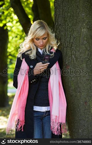 young blond woman with hairstyle in winter dress using her telephone near a tree in a forest