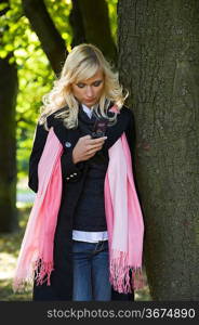 young blond woman with hairstyle in winter dress using her telephone near a tree in a forest