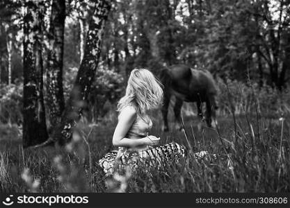 Young blond woman with long hair sitting looking back at a grazing horse on the forest background. Black and white side view, selective focus.. Blonde Woman In The Forest