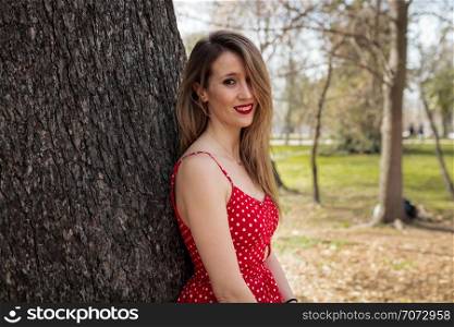 Young blond woman with red dress leaning against a tree