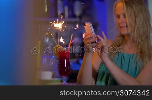 Young blond woman with smart phone taking picture of cocktail with sparkler and then making selfie with it. When fire going down she having a drink