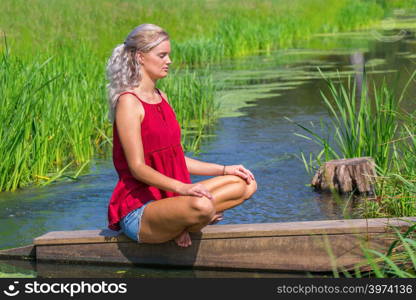 Young blonde caucasian woman meditates at river in nature