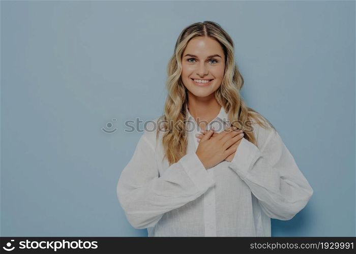 Young blonde female dressed in white shirt relieved with hands resting on her chest, standing relaxed and smiling, being grateful to whoever helped her, isolated in front of light blue background. Relieved blonde woman with hands on her chest