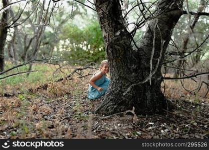 Young blonde girl crouched on the ground near a tree wearing a dress in the sunset field