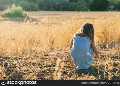 Young blonde girl crouched on the ground playing with the plants wearing a dress in the sunset field