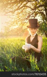 young blonde girl in a brown vintage dress and top hat reads a book in spring garden