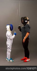 Young blonde girl in a dark coaching outfit and a little boy in a white sports outfit practicing rapier fencing on a gray background. two fencing workout