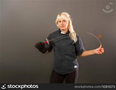 Young blonde girl in a fencing coaching dark uniform stands with a rapier on a gray background. strong fencing girl