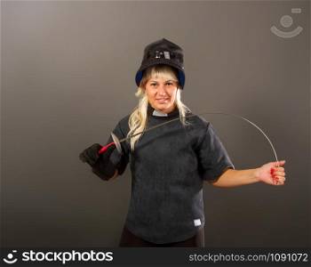 Young blonde girl in a fencing trainer dark uniform and black helmet mask stands with a rapier on a gray background. strong fencing girl