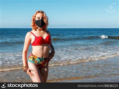 young blonde girl in a red swimsuit and black protective mask standing on the seashore on sunny summer day closeup