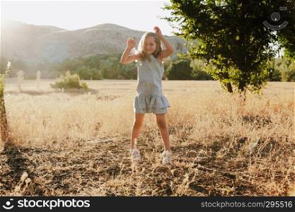Young blonde girl jumping in the field wearing a dress with hills in the background in the sunset